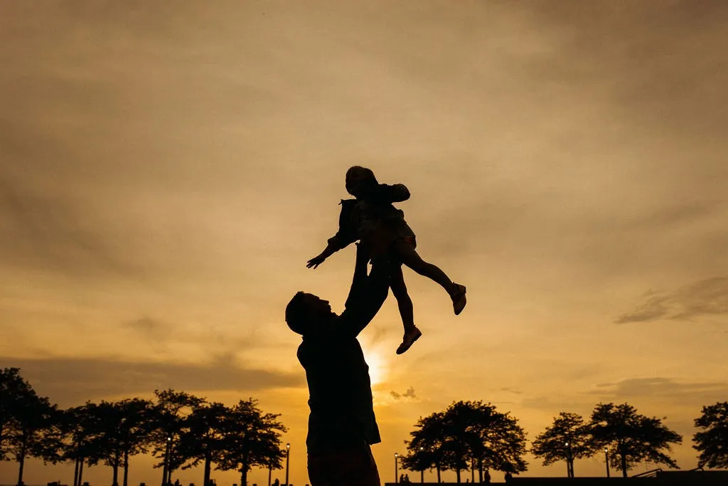 A man lifting a child into the air, silhouetted against a sunset over trees